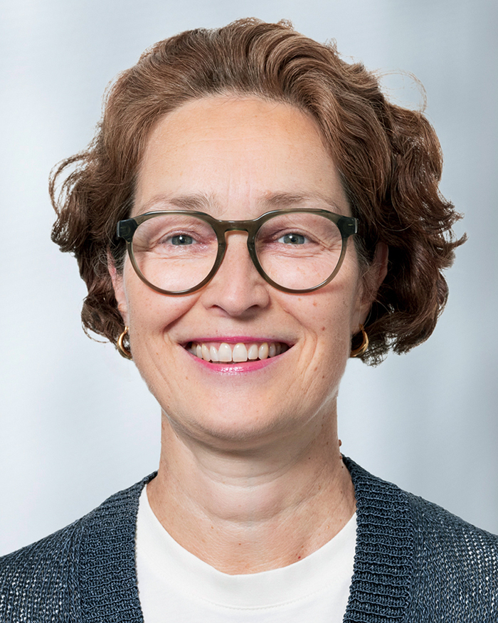 Portrait Tanja Neve-Seyfarth, weiblich lesbare, weisse Person mit mittellangem, lockigem Haar, Brille, Ohr-ringen, weissem Oberteil und grauer Jacke