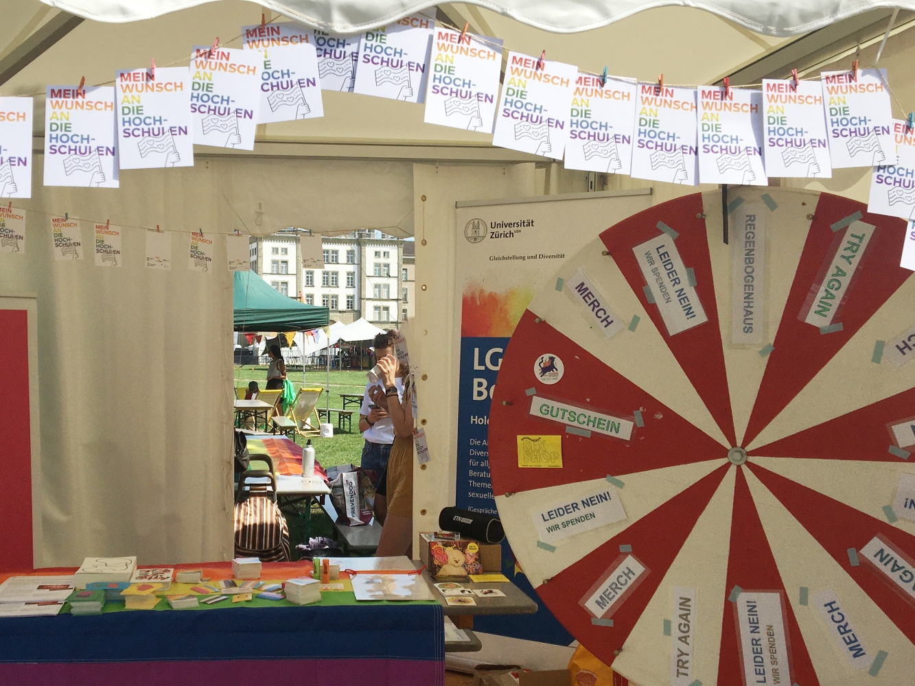 a decorated market stall on the topic of equality and diversity at universities, many materials are laid out on the table of the stall, roll ups are in the stall, in front of the stall there is a red and white striped wheel of fortune