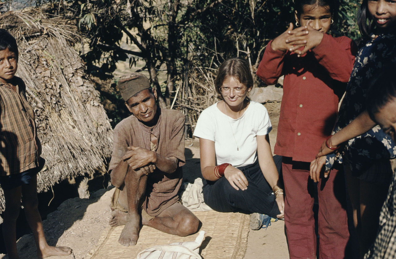 Ulrike Müller-Böker in Nepal, eine junge, weisse Frau sitzt neben einem farbigen Mann auf dem Boden, um die beiden herum stehen einheimische Kinder